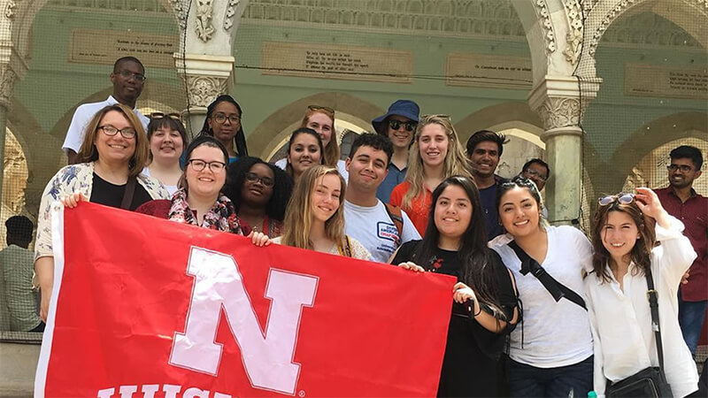 Students hold University of Nebraska-Lincoln flag while posing in front of Intricately carved pillars.